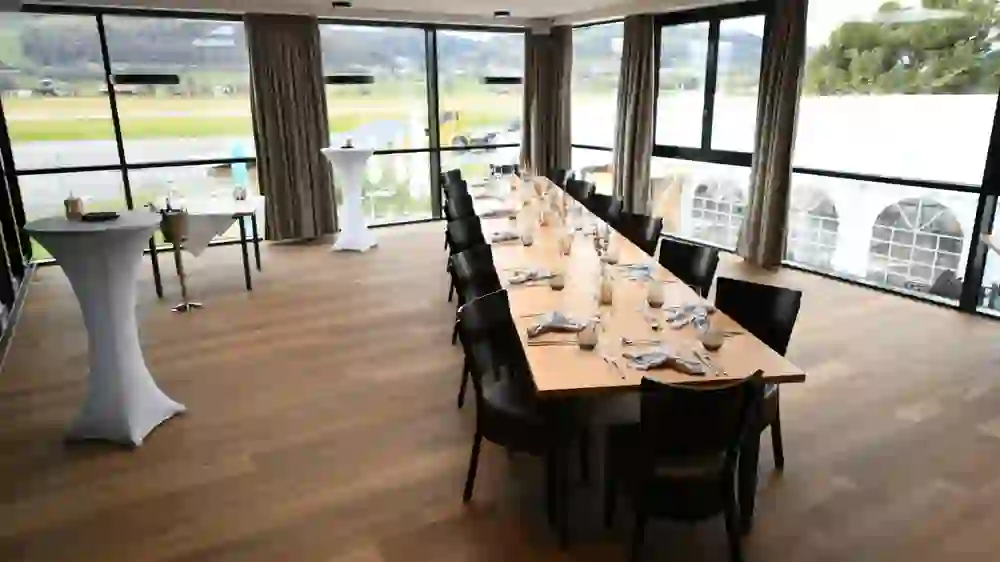 L'immagine mostra un lungo tavolo da pranzo in una stanza con finestre panoramiche che si affacciano su una pista. La tavola è imbandita per un pasto e ci sono tavoli da bar con refrigeratori per lo champagne. Si tratta di un ambiente elegante, probabilmente in un hotel o in un ristorante dell'aeroporto.