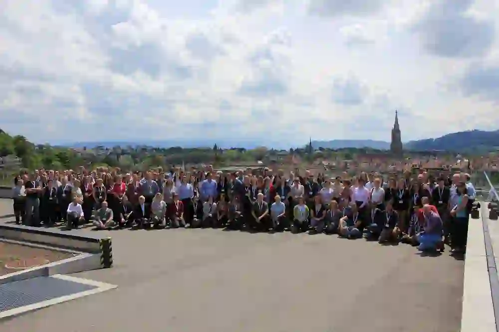 Auf dem Foto ist eine große Gruppe von Menschen, die für ein Gruppenbild posieren, vor einer städtischen Kulisse mit Blick auf Bern, der Hauptstadt der Schweiz.