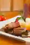 Plat gastronomique composé d'un steak, d'un produit enveloppé de lard, de croquettes, de champignons et de garnitures sur une assiette blanche. En arrière-plan, des tomates cerises et des oignons.