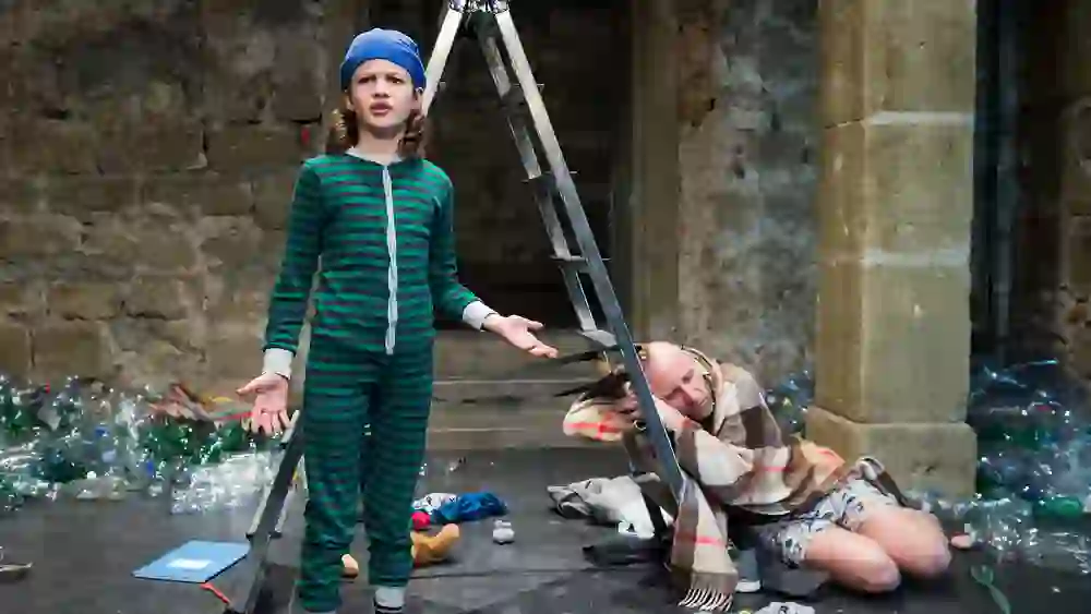 A child in striped pyjamas stands confused next to a ladder, while an exhausted-looking person lies next to it, wrapped in a blanket, surrounded by clutter and plastic rubbish, creating a thought-provoking, perhaps even critical scene.