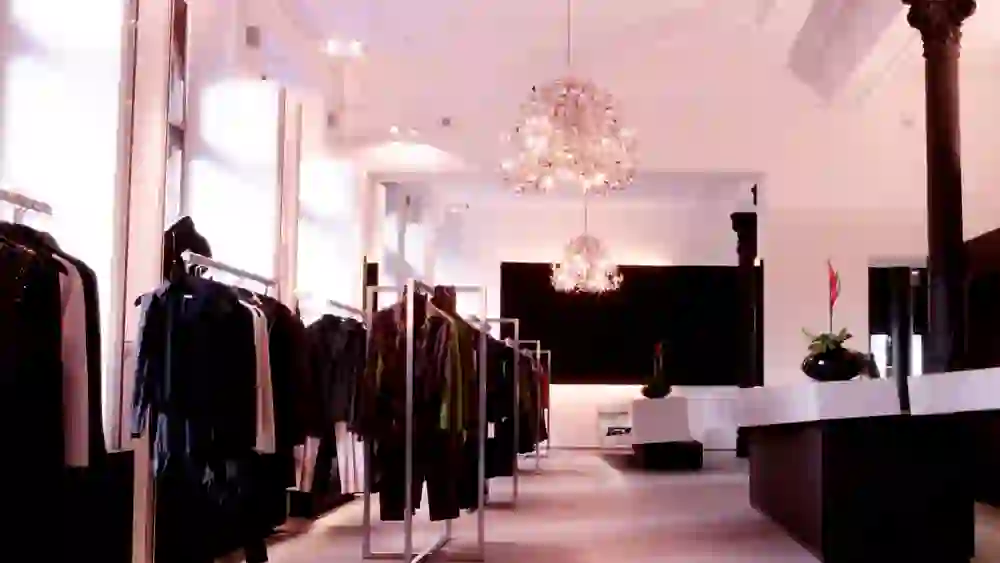 Auf diesem Foto ist der Innenraum von Aerni Haar Kleid Bar Spa zu sehen. Die Beleuchtung durch funkelnde Kristalllüster verleiht dem Raum eine anspruchsvolle und warme Ausstrahlung. Die Bekleidungsstücke sind auf Ständern präsentiert, die eine geradlinige, übersichtliche Einkaufsatmosphäre schaffen. In der Mitte sorgen dunkel gefärbte Säulen und schwarzglänzende Akzente für einen dramatischen Kontrast zur hellen Umgebung und unterstreichen das elegante Ambiente des Geschäfts.