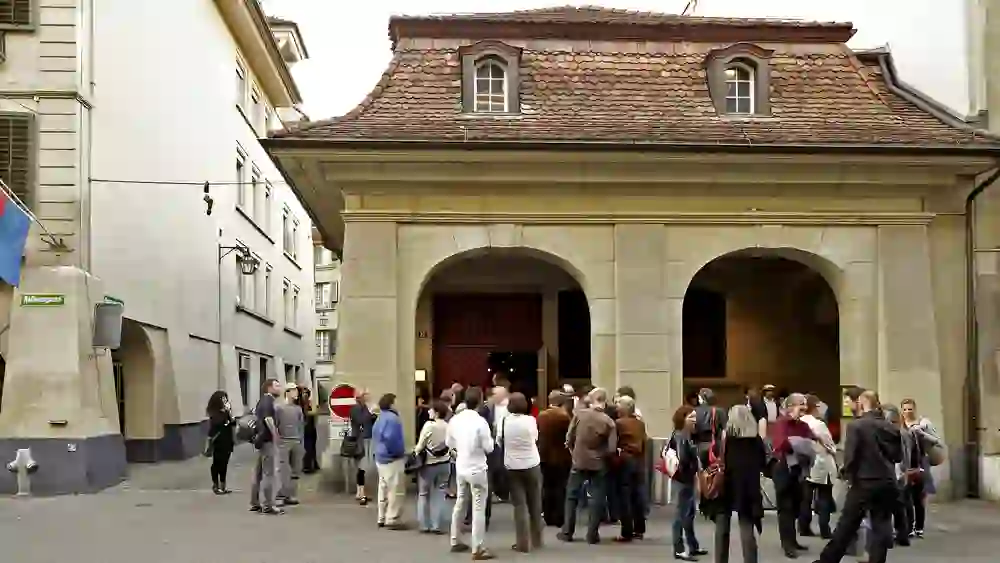 Vor dem Schlachthaus Theater in Bern versammelt sich eine Gruppe von Menschen in angeregter Unterhaltung, voller Erwartung auf die kommende Vorstellung. Die Stimmung ist gesellig und erwartungsvoll.