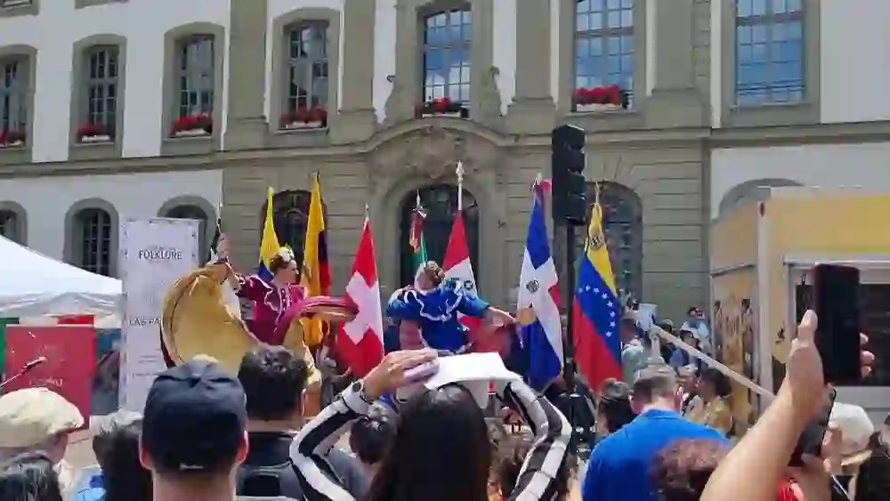 La photo montre une démonstration énergique de danses traditionnelles mexicaines lors d'un festival. Des danseurs en costumes vifs balancent leurs jupes devant une foule enthousiaste, encadrée par des drapeaux internationaux et une architecture historique.