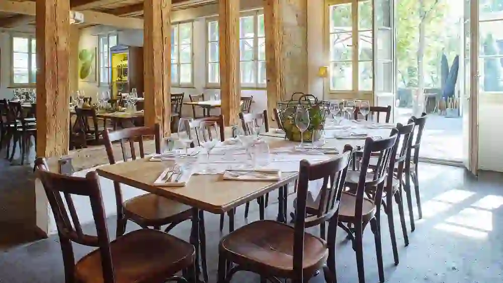 Das Bild zeigt ein helles, luftiges Restaurant mit natürlichen Holzbalken und großen Fenstern, die für viel Tageslicht sorgen. Elegante Tischdecken und gepflegtes Tafelsilber laden zu einem gehobenen Essen ein.