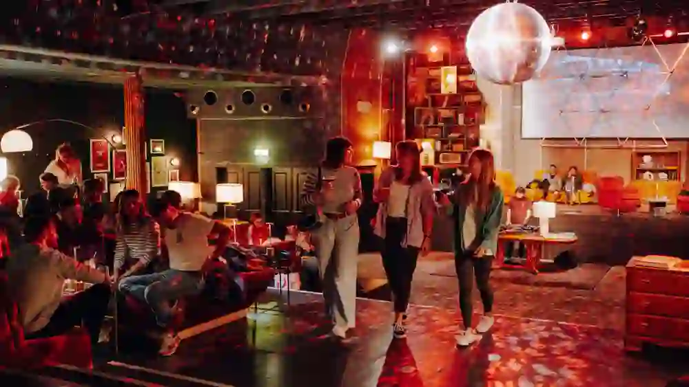 Das Bild zeigt eine lebhafte Szene in einer gemütlichen, gut beleuchteten Bar oder Lounge. Im Vordergrund sind mehrere Personen zu sehen, die sich unterhalten und Getränke in der Hand halten. Im Hintergrund gibt es weitere Sitzgelegenheiten, dekorative Lampen und eine große Projektionsfläche, auf der ein Bild angezeigt wird. An der Decke hängt eine Discokugel, die für bunte Lichtreflexe im Raum sorgt. Die Atmosphäre wirkt entspannt und einladend.