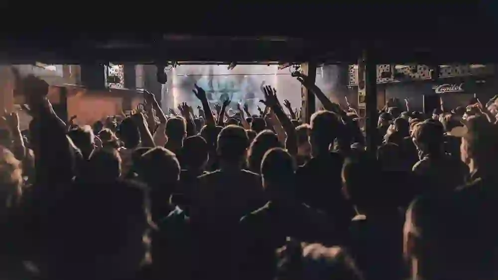 Das Foto fängt die energetische Stimmung eines Konzertes im Old Capitol ein, mit einer Menschenmenge, die begeistert die Hände in die Höhe streckt. Die Beleuchtung schafft eine fesselnde Atmosphäre, und die Silhouetten der Zuschauer deuten auf eine Nacht voller Musik und Tanz hin.