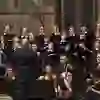 The picture shows a choir and an orchestra during a concert. The conductor is leading the group, which is singing and playing intensively. The scene seems to take place in a room with wall paintings that has a historical feel.
