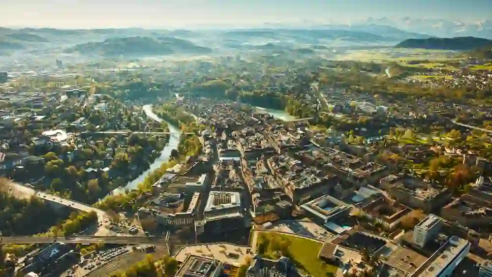 Eine Luftaufnahme der Stadt Bern zeigt die dichte Bebauung, die Aare, die sich durch die Stadt schlängelt, umgeben von Grünflächen und dem Alpenpanorama im Hintergrund.