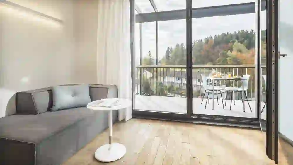 Ein gemütliches Wohnzimmer im b20 serviced apartmens bern mit Sofa und großem Fenster, das zu einer Terrasse mit Herbstblick führt, strahlt Ruhe und Komfort aus und lädt zum Entspannen in einer hellen, freundlichen Umgebung ein.