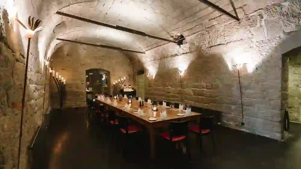 Dans une cave voûtée historique, on peut voir une table festive, entourée de murs en pierre et de lampes torches d'ambiance, qui créent une atmosphère intime et rustique. Idéal pour un dîner spécial.
