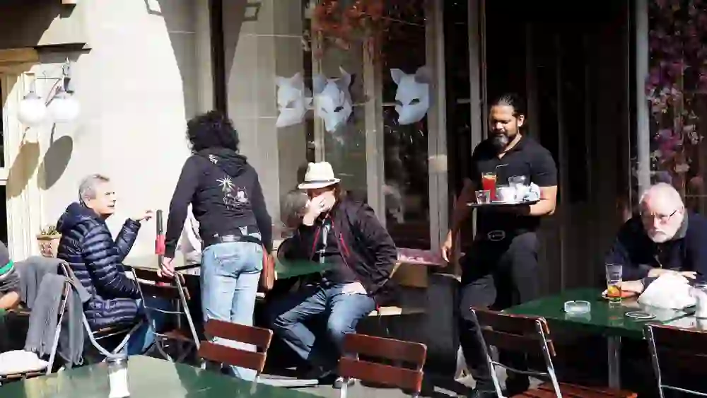 Sur la photo, on peut voir une scène animée au Café des Pyrénées à Berne. Un serveur apporte des boissons tandis que des clients sont assis à des tables en plein air et discutent. Une personne portant un chapeau clair semble tenir une conversation, tandis qu'une autre a l'air pensif. En arrière-plan, on distingue le café avec des fenêtres sur lesquelles on peut voir des représentations artistiques de têtes d'ours et des décorations fleuries. L'ambiance est détendue et conviviale.