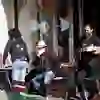 L'immagine mostra una scena vivace al Café des Pyrénées di Berna. Un cameriere porta le bevande mentre gli ospiti sono seduti ai tavoli all'aperto a chiacchierare. Una persona con un cappello chiaro sembra conversare, mentre un'altra ha un'aria pensierosa. Sullo sfondo, il caffè è caratterizzato da finestre con raffigurazioni artistiche di teste di orso e decorazioni floreali. L'atmosfera è rilassata e socievole.
