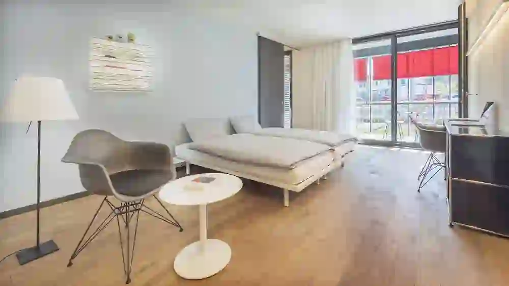 Ein modernes, minimalistisch eingerichtetes Zimmer im b20 serviced apartmens bern mit natürlichen Holzelementen, das eine entspannte und saubere Atmosphäre bietet. Die großen Fenster lassen viel Licht herein und schaffen ein Gefühl von Offenheit und Ruhe.