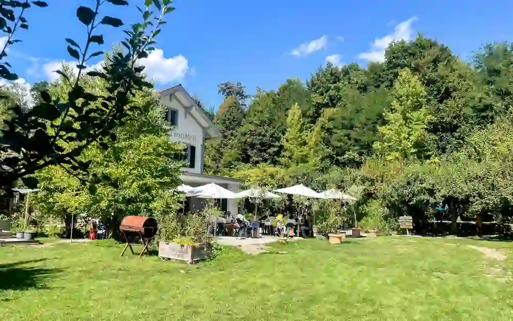 Sur la photo, on peut voir un jardin-restaurant idyllique. Des parasols offrent de l'ombre aux clients qui se détendent dehors par un temps agréable. L'ambiance semble calme et sereine, un sentiment de sérénité et de plaisir des loisirs imprègne la scène.