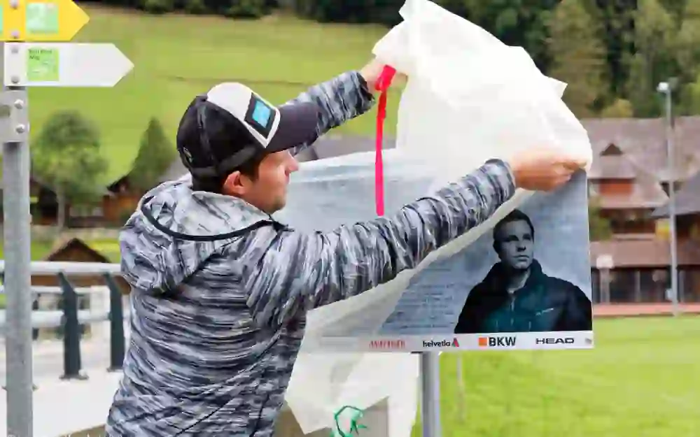 Das Bild zeigt einen Mann, der aktiv ein Poster an einem Wegweiser befestigt, der einen Wanderweg kennzeichnet. Er wirkt konzentriert und engagiert bei seiner Tätigkeit, was eine Atmosphäre von Tatendrang und sorgfältiger Vorbereitung in einer idyllischen, ländlichen Umgebung schafft.