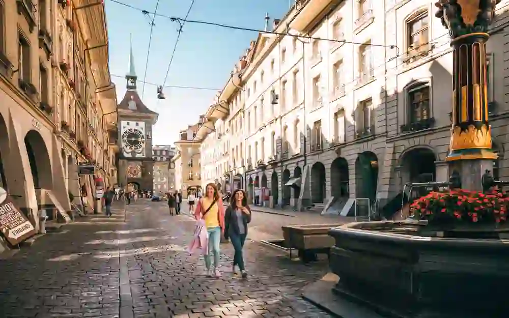 L'immagine mostra due donne allegre che passeggiano nel vivace e soleggiato centro storico di Berna, con la famosa torre Zytglogge sullo sfondo. L'ambiente circostante emana un'atmosfera rilassata e invitante.