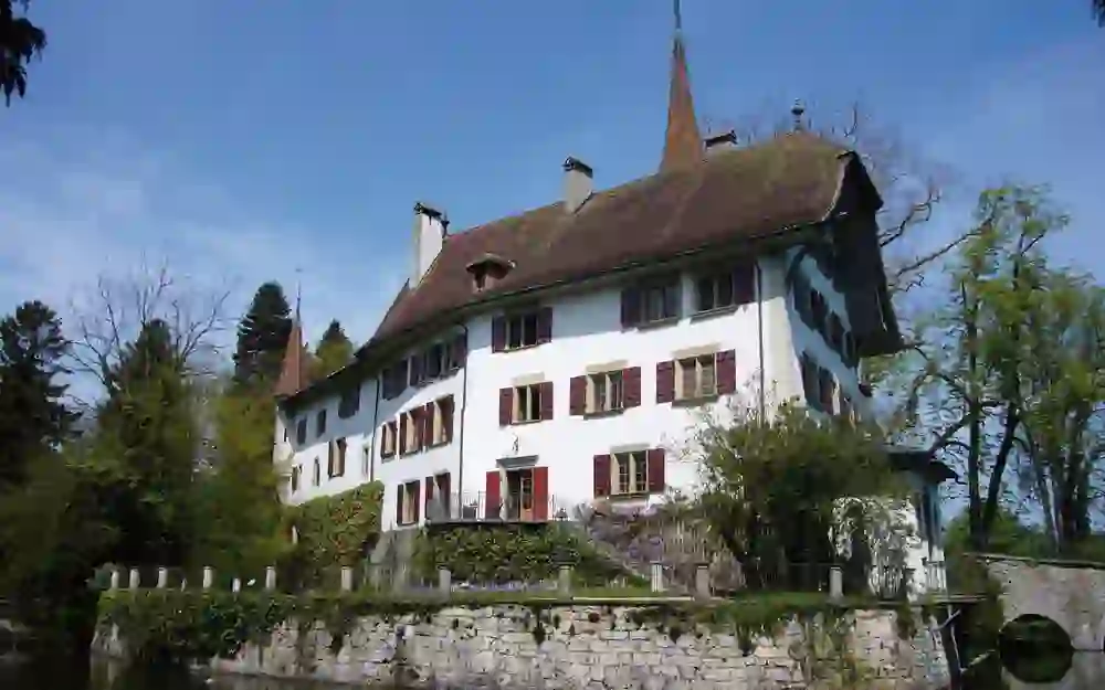 Das Foto zeigt das Schloss Landshut in einer ländlichen Szenerie. Es steht am Ufer eines stillen Gewässers, welches das Gebäude und die umgebende Vegetation sanft reflektiert.
