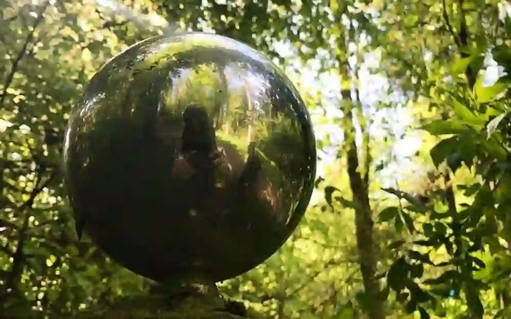 Das Bild zeigt eine glänzende, kugelförmige Skulptur in einem Wald, die die Umgebung reflektiert und eine verschwommene Figur im Hintergrund zeigt. Die Stimmung ist geheimnisvoll und einladend, inspiriert zur Reflexion und vermittelt eine ruhige Verbindung zur Natur.