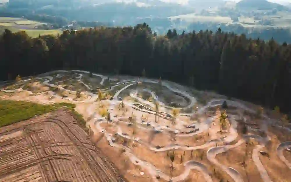 The picture shows an aerial view of a pump track in a rural landscape, surrounded by fields and a forest.