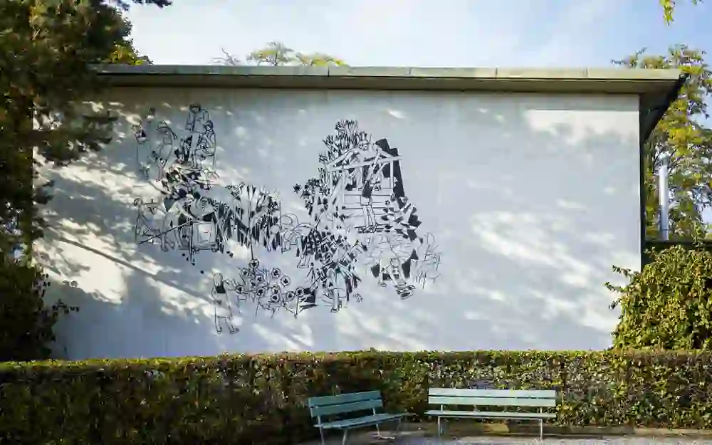 Auf dem Bild ist eine Wand mit einem großflächigen, modernen Wandgemälde zu sehen, das lebhafte und abstrakte Figuren darstellt. Zwei Bänke vor der Wand laden zum Verweilen und Betrachten ein, was eine ruhige und nachdenkliche Stimmung schafft. Die Umgebung wirkt einladend und künstlerisch.