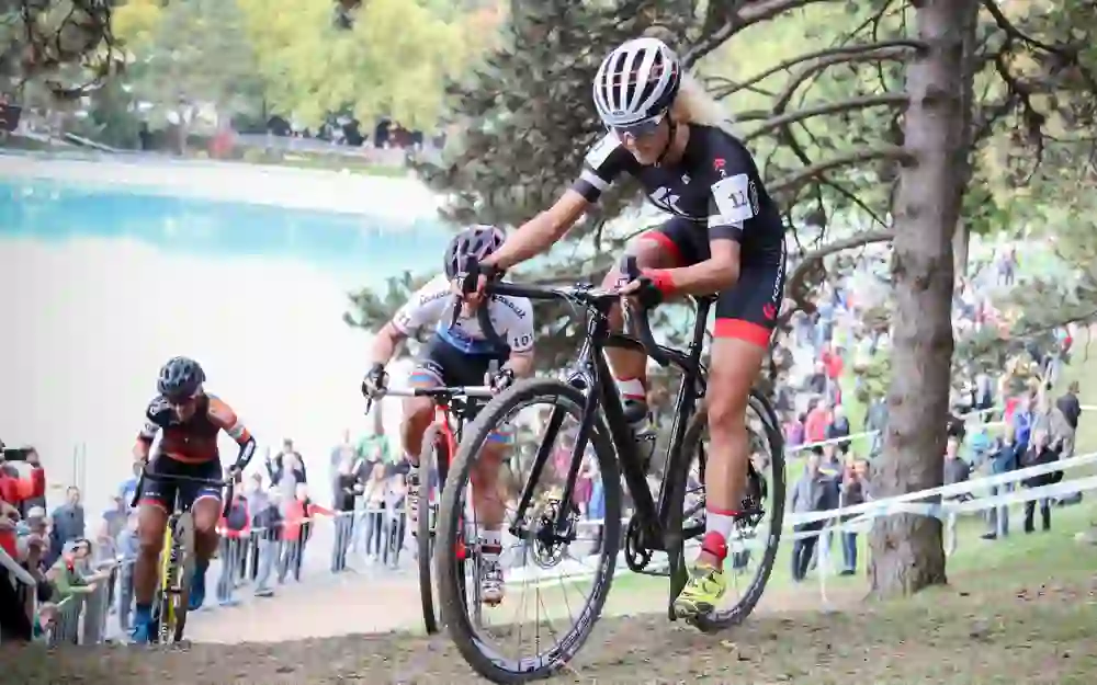 Radsportlerinnen konkurrieren mit Anstrengung und Fokus bei einem Cyclocross-Rennen, unterstützt von den Anfeuerungen des Publikums im Hintergrund – eine Szene voller Energie und sportlicher Leidenschaft.