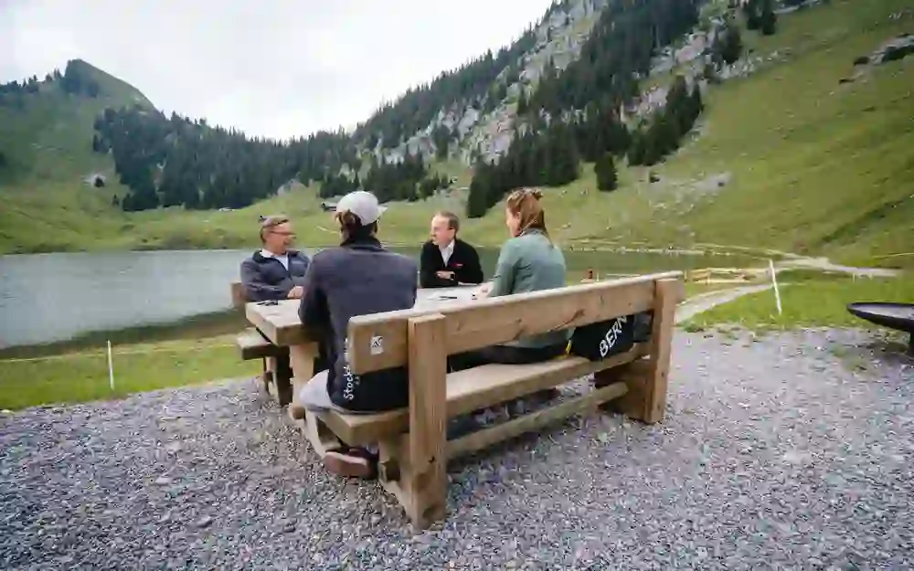Eine Gruppe sitzt an einem Holztisch in einer alpinen Landschaft, umgeben von Bergen und einem See. Sie scheinen in ein angeregtes Gespräch vertieft zu sein, das die friedliche und erhabene Naturkulisse widerspiegelt.