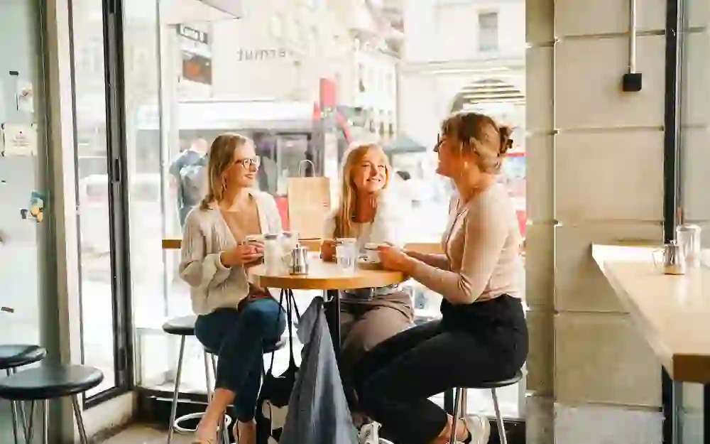 Drei Freundinnen sitzen lächelnd an einem Café-Tisch, genießen Getränke und eine lebhafte Unterhaltung. Sonnenlicht füllt den Raum, welcher eine warme und einladende Atmosphäre schafft.