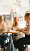 Three friends sit smiling at a café table, enjoying drinks and lively conversation. Sunlight fills the room, creating a warm and inviting atmosphere.
