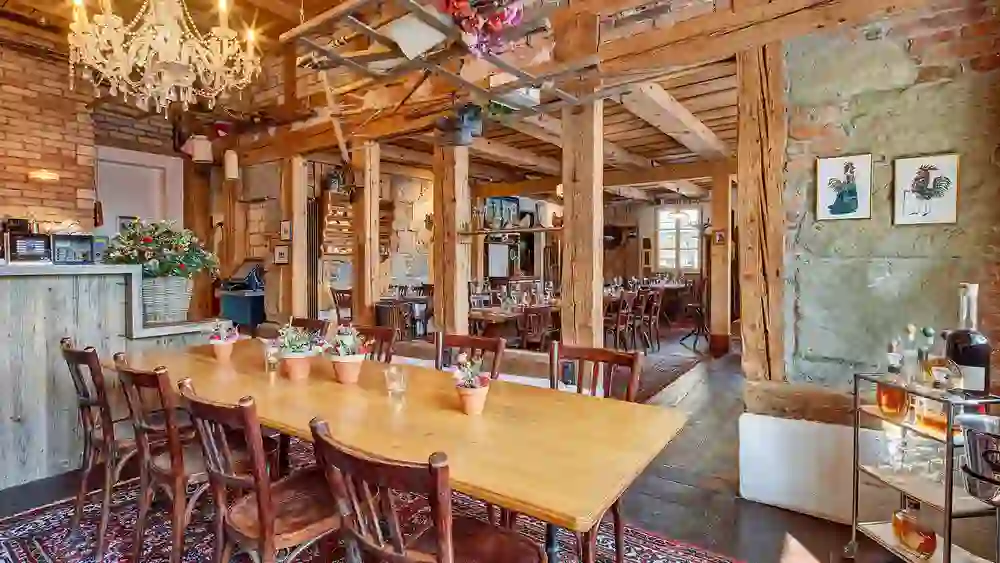 Das Foto zeigt ein einladendes Restaurant mit rustikaler Einrichtung, Holzmöbeln und einem großen Kronleuchter. Blumen auf den Tischen und ein Orientteppich verleihen dem Raum eine warme Atmosphäre.