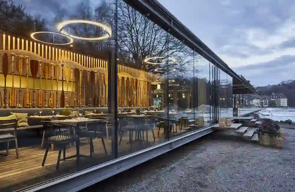 The photo shows the interior of a busy restaurant at dusk with a view of an illuminated outdoor area and the flowing River Aare. The warm interior lighting and outdoor string lights give the place a cosy and inviting atmosphere. Guests can dine inside or enjoy the view from the terrace. The scene conveys a sense of relaxation and socialising in a picturesque setting.