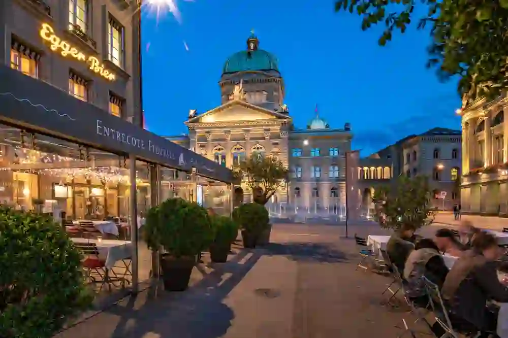 The picture shows a restaurant at dusk with guests sitting outside. A magnificent building can be seen in the background. The lighting of the restaurant and the blue hour create a calm and pleasant atmosphere, ideal for an outdoor dinner.