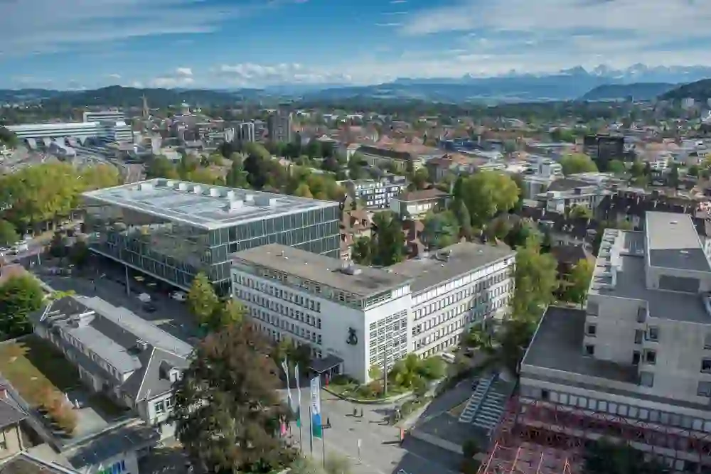 Ein Blick auf das moderne Stadtviertel von Bern mit einem Vordergrund aus Glasgebäuden und einem Hintergrund, der die majestätischen Alpen zeigt, vermittelt eine Atmosphäre von Fortschritt und der Harmonie von Stadt und Natur.