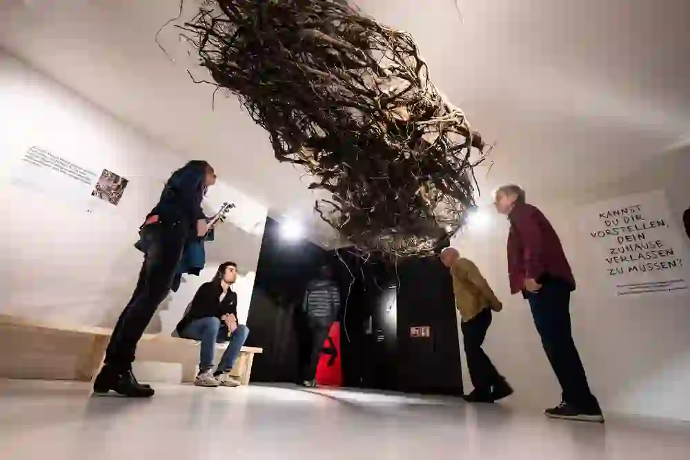 Sur l'image, on voit un groupe de personnes debout dans une pièce avec une installation artistique. Au-dessus d'eux, une grande motte de racines est suspendue au plafond, ce qui crée un fort impact visuel. Les personnes contemplent l'art ou discutent, tandis qu'un panneau sur le mur pose une question qui incite à la réflexion. L'ambiance est à la réflexion et à l'interactivité.