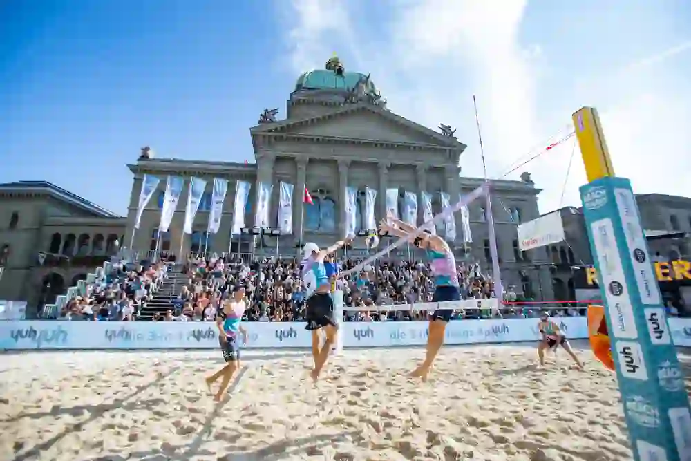 Auf dem Bild ist ein Beachvolleyballspiel vor einer beeindruckenden historischen Kulisse zu sehen. Die Zuschauertribünen sind voll besetzt, während zwei Spieler gerade den Ball am Netz blocken. Im Hintergrund steht ein großes Gebäude mit der Aufschrift "Curia Confederationis Helveticae". Es handelt sich um ein Event der Yuh Beach Tour, erkennbar an den Bannern und Plakaten.