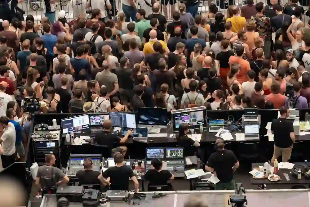 Das Bild zeigt eine große Menschenmenge bei einem Event, fokussiert auf das Geschehen. Im Vordergrund arbeiten Techniker intensiv an Steuerpulten, umgeben von Monitoren und Ausrüstung, was eine konzentrierte Atmosphäre schafft.