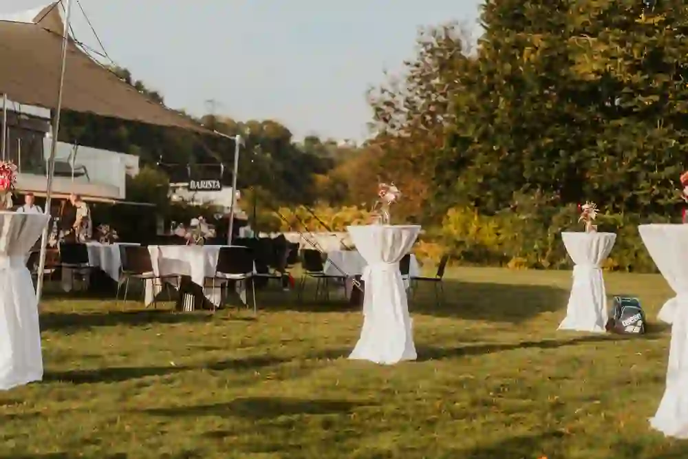 Allestimento estivo con tavoli da bar bianchi e sottili decorazioni floreali sul prato, tenda BARISTA sullo sfondo.