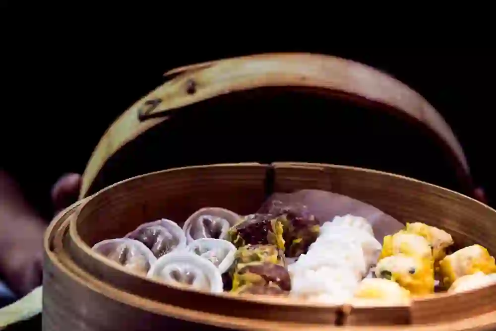 L'image montre une étuve en bambou remplie de différents types de dim sum. Les raviolis et bouchées cuits à la vapeur de différentes couleurs sont appétissants et accueillants. L'atmosphère chaleureuse et conviviale invite à une expérience culinaire.