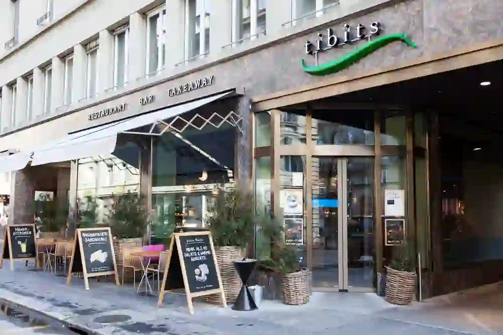 The picture shows the exterior of the "tibits" restaurant in Bern with an awning and a green logo in the shape of a chilli pepper above the entrance. In front of it are tables and chairs for guests outside, surrounded by advertising boards and plants in baskets. It appears to be a modern, inviting restaurant that also offers takeaway, as indicated on the sign above the entrance.