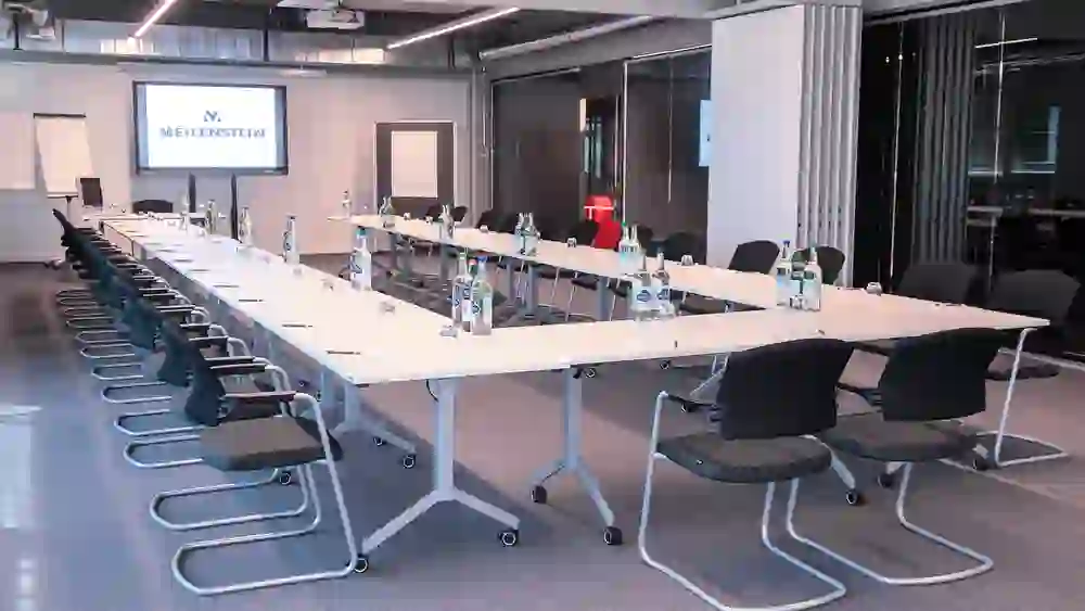Das Bild zeigt einen modernen, hellen Seminarraum, ausgestattet mit einer U-förmigen Tischanordnung, Stühlen und Präsentationstechnik, bereit für eine informative und interaktive Tagung oder Schulung.