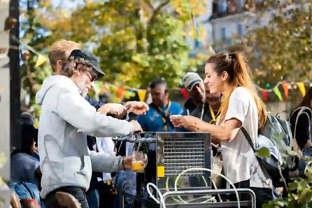 Eine ausgelassene Szene auf einem Festival: Ein Mann schenkt Bier ein, während eine lächelnde Frau wartet, umgeben von fröhlichen Menschen an einem sonnigen Tag mit bunten Girlanden im Hintergrund.