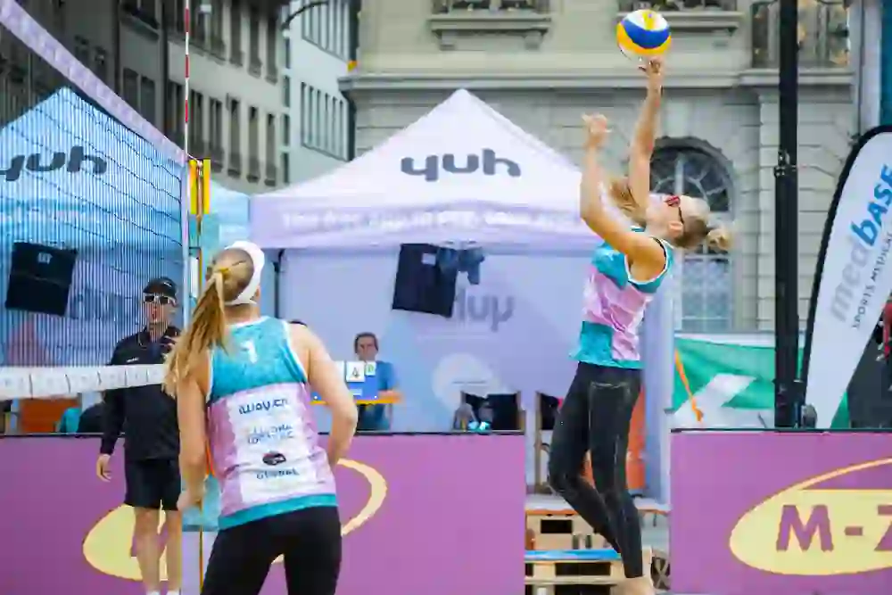 L'immagine mostra una partita di beach volley. Una giocatrice salta per colpire la palla mentre la sua compagna di squadra, entrambe in maglietta turchese, si trova a rete. Sullo sfondo ci sono striscioni e tende di sponsor, tra cui "Yuh" e "medbase", e un arbitro. La scena fa pensare a un torneo professionale.