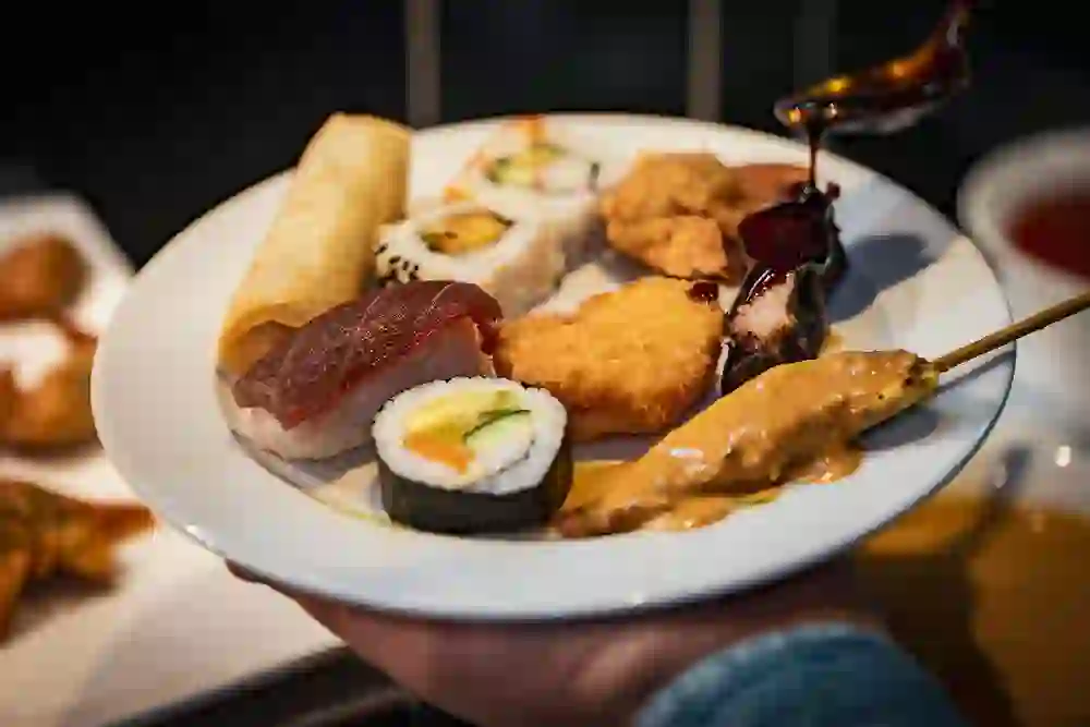 L'image montre une assiette avec une sélection de plats asiatiques, dont des sushis, des rouleaux de printemps, du poulet frit et des brochettes de viande en sauce. Les multiples arômes et textures de l'assiette apportent une touche de luxe culinaire et de plaisir, parfaite pour les amateurs de cuisine asiatique.