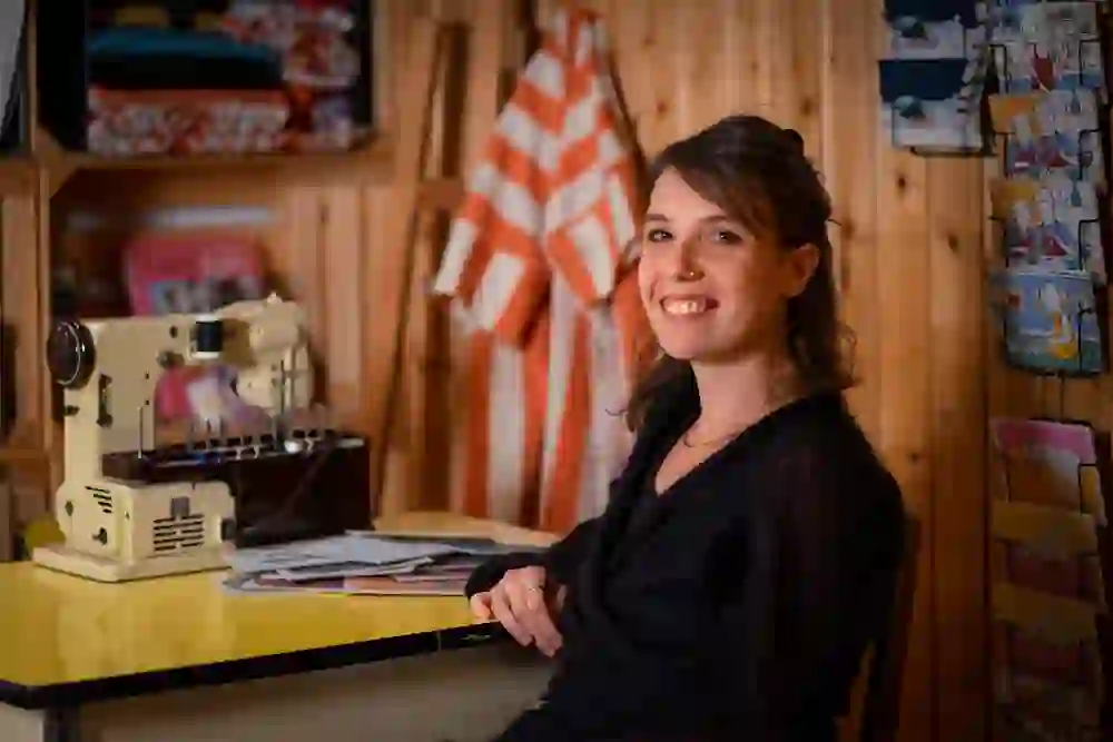 The photo shows Nora Ryser, a young woman sitting next to an old sewing machine. Her smile and the cosy, wood-panelled room with colourful fabrics and postcards radiate warmth and creativity.