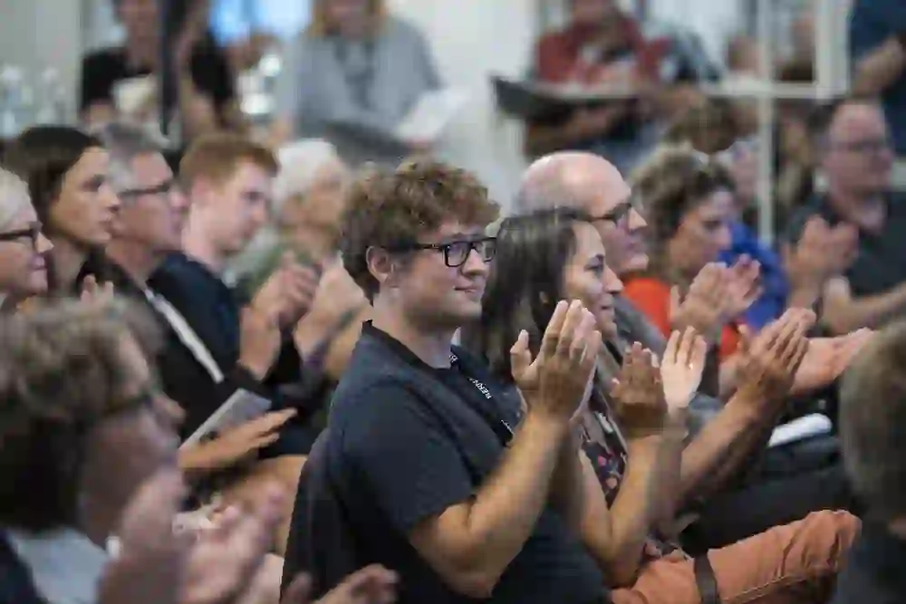 Ein Publikum klatscht Beifall, eingetaucht in die zufriedene Stimmung einer Veranstaltung. Im Zentrum ein junger Mann, der mit einem Lächeln die Aufführung anerkennend würdigt.