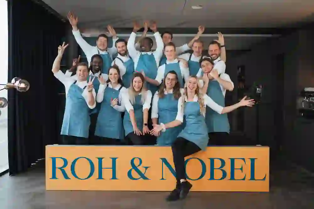Il team di Roh & Nobel posa allegramente per una foto di gruppo davanti alla lavagna con il logo arancione.