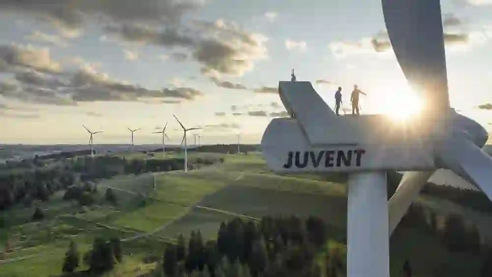 Das Bild zeigt ein Windkraftwerk mit der Aufschrift „JUVENT“ in einer ländlichen Landschaft bei Sonnenuntergang. Zwei Personen stehen oben auf dem Windrad, was auf Wartungsarbeiten hinweist.