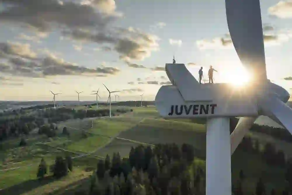 L'immagine mostra una turbina eolica con la scritta "JUVENT" in un paesaggio rurale al tramonto. Due persone sono in piedi in cima alla turbina eolica, a indicare lavori di manutenzione.