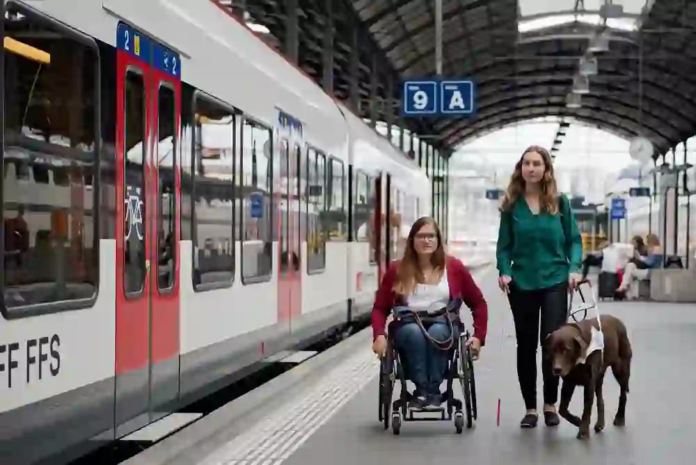 L'image montre deux personnes dans une gare, l'une en fauteuil roulant et l'autre malvoyante avec un chien guide d'aveugle. Elles semblent plongées dans une interaction alors qu'elles se trouvent à côté d'un train. L'ambiance semble sereine et connectée.
