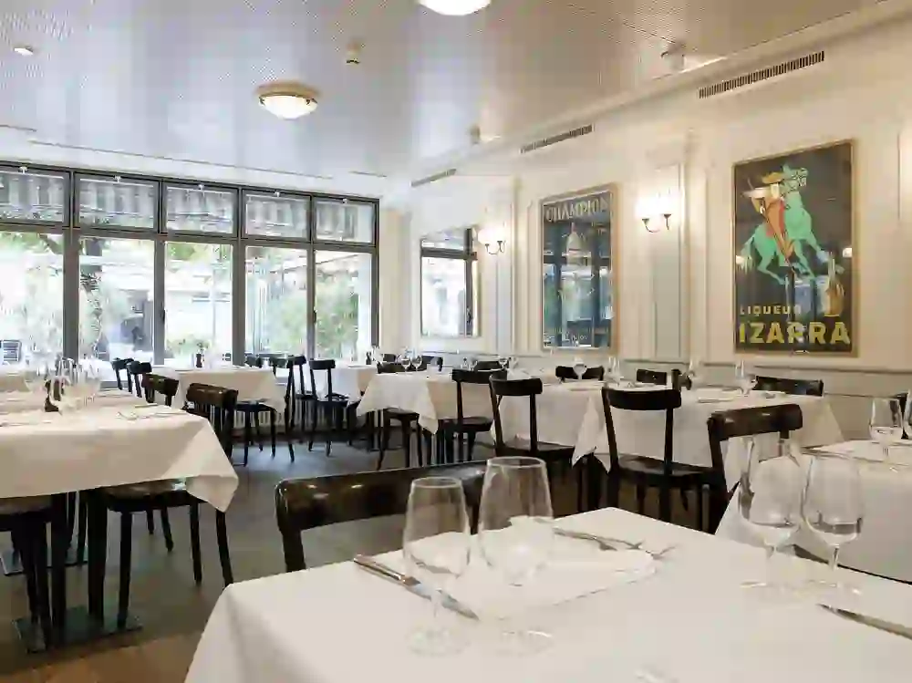 Das Bild zeigt das elegante Interieur eines Restaurants mit gedeckten Tischen, weißen Tischdecken und Weingläsern. An den Wänden hängen Vintage-Poster. Große Fenster bieten Blick auf einen Außenbereich. Die Stimmung wirkt ruhig und einladend, bereit für die Ankunft von Gästen.
