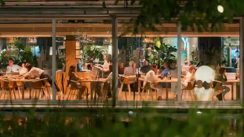 Das Bild zeigt eine sonnige Terrasse eines eleganten Gebäudes mit klassischer Fassade. Die gedeckten Tische und großen Sonnenschirme laden zu entspannten Stunden im Freien ein, während die farbenfrohe Blumenpracht eine angenehme, freundliche Atmosphäre schafft.